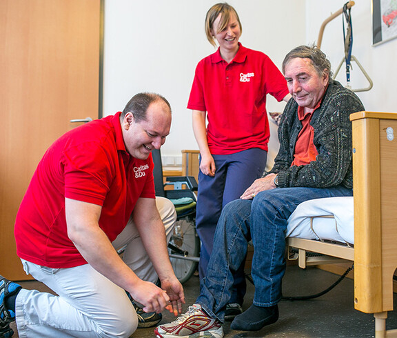 Zwei BetreuerInnen der Caritas Altenpension helfen einem Mann beim Schuhe anziehen.