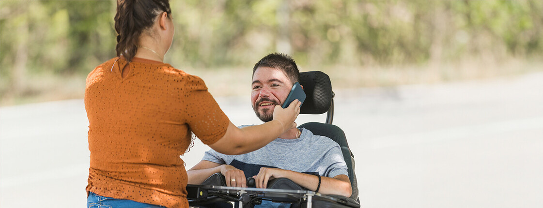  Eine Frau hält einem lächelnden Mann im Rollstuhl ein Smartphone ans Ohr. Sie befinden sich draußen vor einem unscharfen grünen Hintergrund.