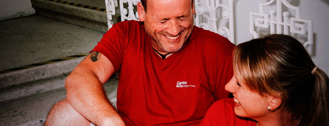 Ein Mann und eine Frau mit roten T-Shirts mit Caritas-Logo sitzen auf den Stiegen und lachen gemeinsam. Die Frau hat einen Aktenordner in der Hand.