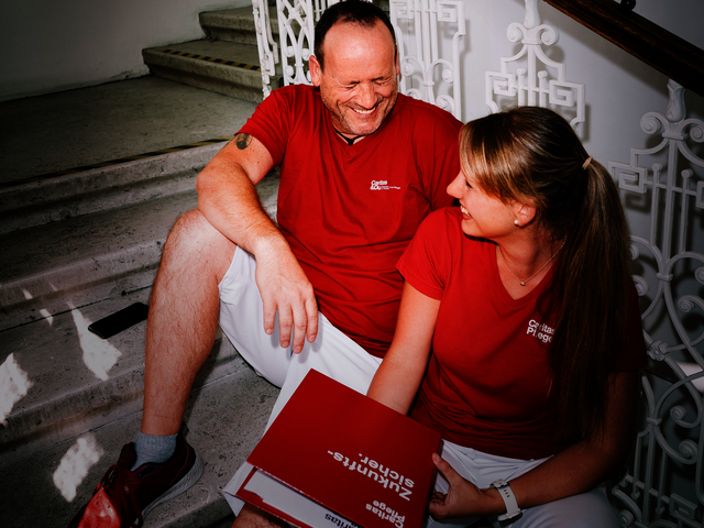 Ein Mann und eine Frau mit roten T-Shirts mit Caritas-Logo sitzen auf den Stiegen und lachen gemeinsam. Die Frau hat einen Aktenordner in der Hand.