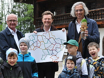 Caritas Direktor Johannes Dines  und der Salzburger Freichlichtmuseums-Direktor stehen mit Kindern auf einem Schotterplatz und halten ein Plakat hoch.